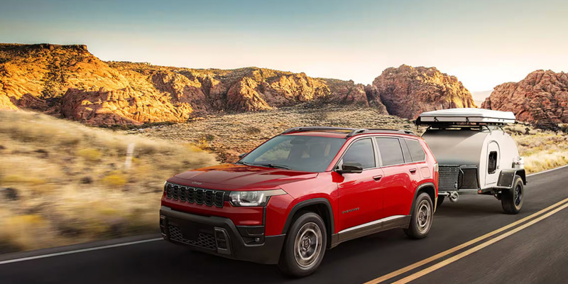 Red Cherokee SUV towing a camper trailer on a desert highway with rocky mountains in the background.