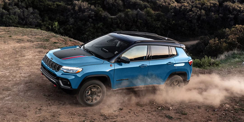 Blue Jeep Compass SUV driving off-road on a dirt trail, kicking up dust while navigating uneven terrain.