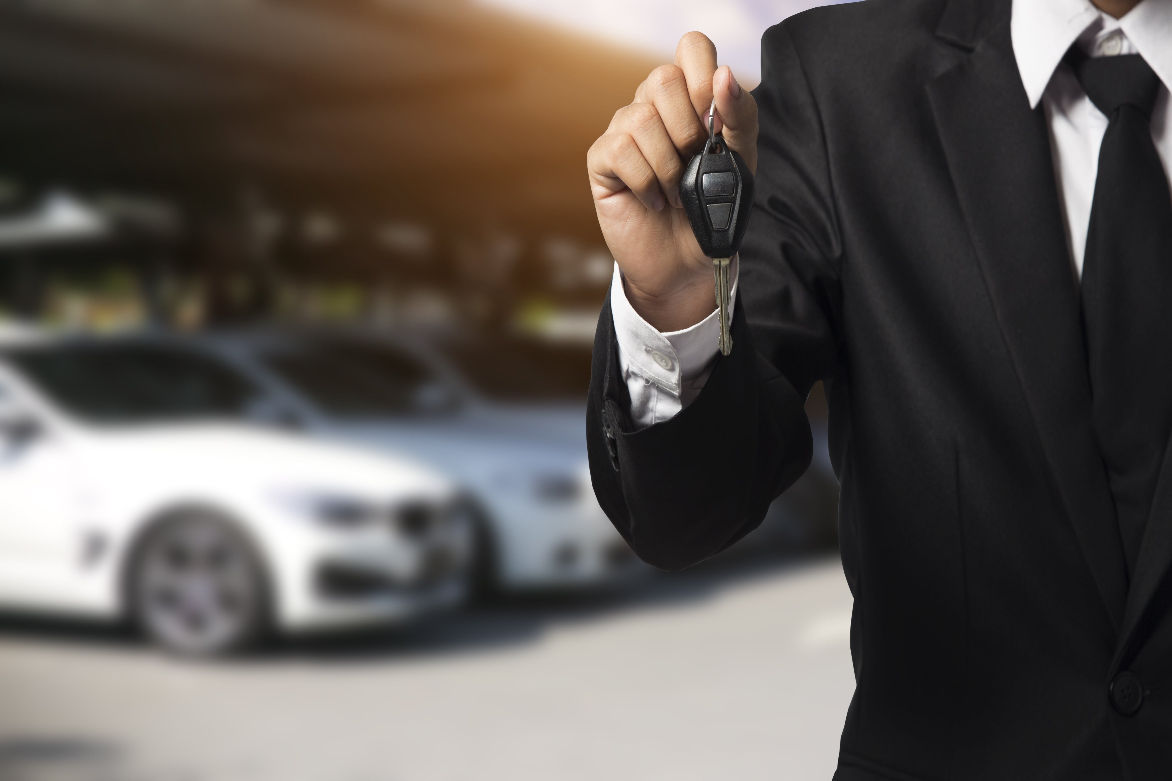 Person in a suit holding car keys with a vehicle dealership lot in the background.